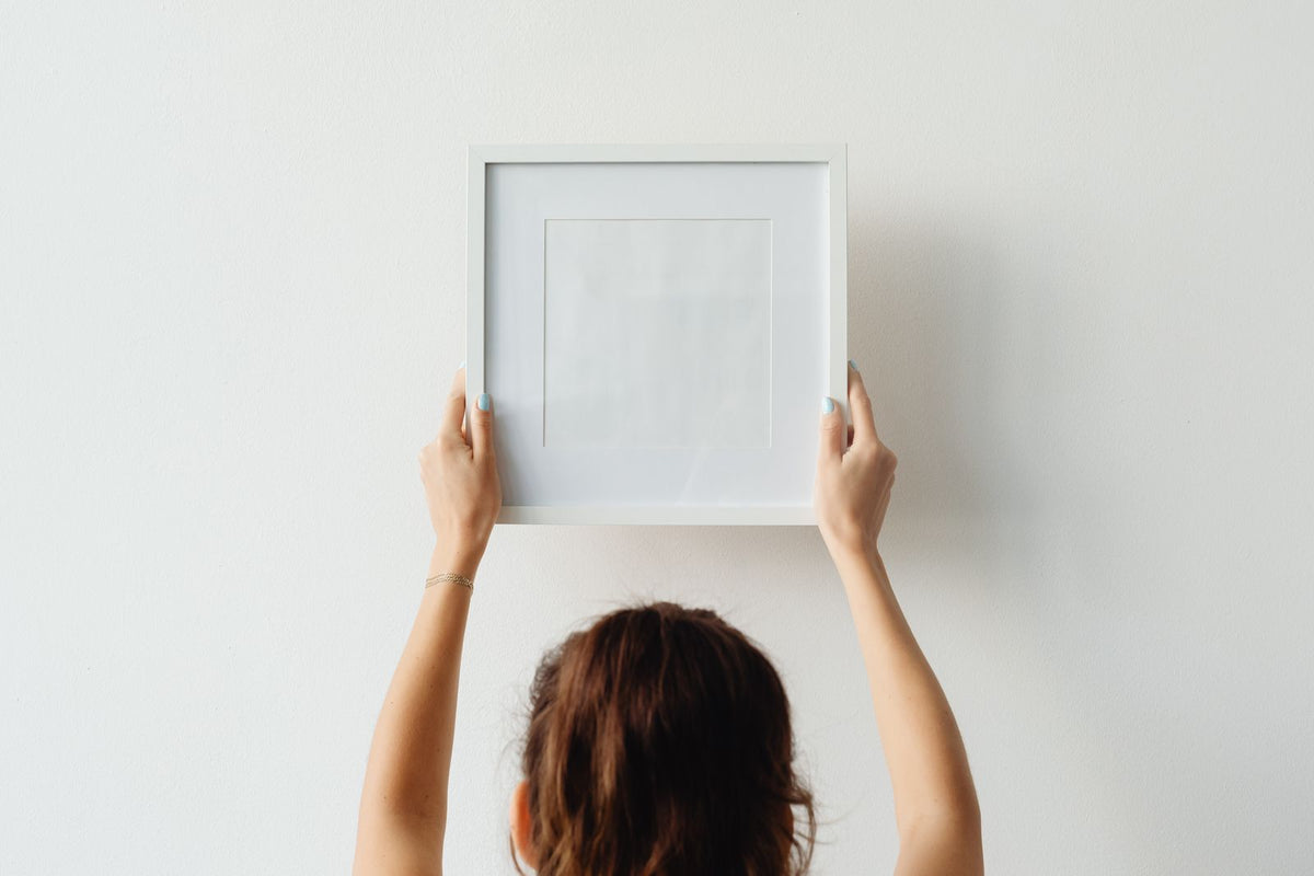 Femme positionnant un cadre sur un mur blanc, à hauteur des yeux